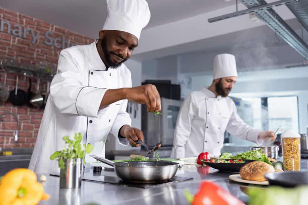 African american chef cooking delicious meals with rosemary or basil in a restaurant kitchen, sprinkles herbs leaves on the stove.