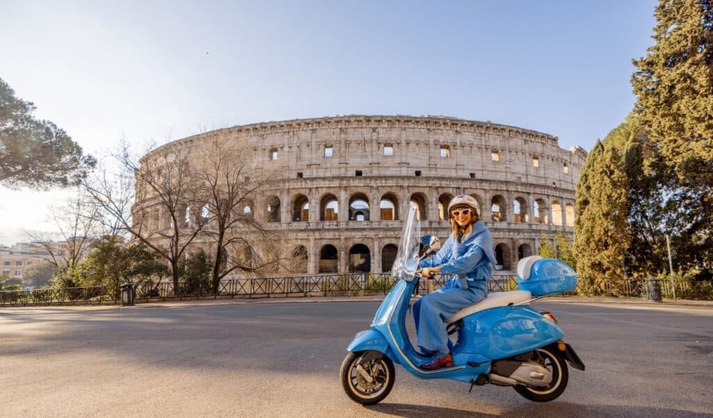 A fashionable woman in a blue outfit rides a classic scooter near the Colosseum in Rome, Italy. The golden sunlight enhances the vibrant city atmosphere, showcasing Italian lifestyle