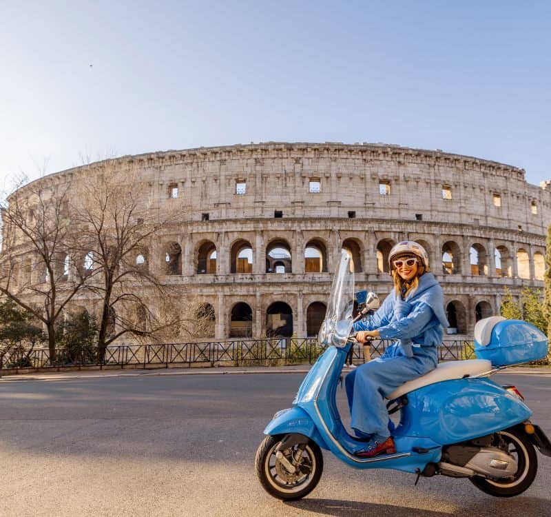 A fashionable woman in a blue outfit rides a classic scooter near the Colosseum in Rome, Italy. The golden sunlight enhances the vibrant city atmosphere, showcasing Italian lifestyle
