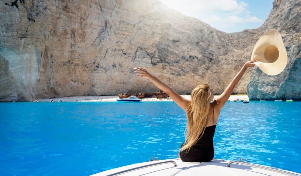 A happy tourist woman sitting on a boat enjoys the beautiful view to the turquoise sea of Shipwreck Navagio Beach, Zakynthos island, Greece