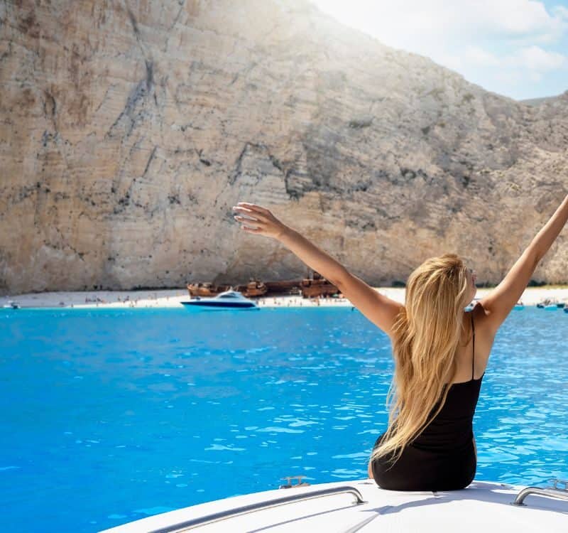A happy tourist woman sitting on a boat enjoys the beautiful view to the turquoise sea of Shipwreck Navagio Beach, Zakynthos island, Greece