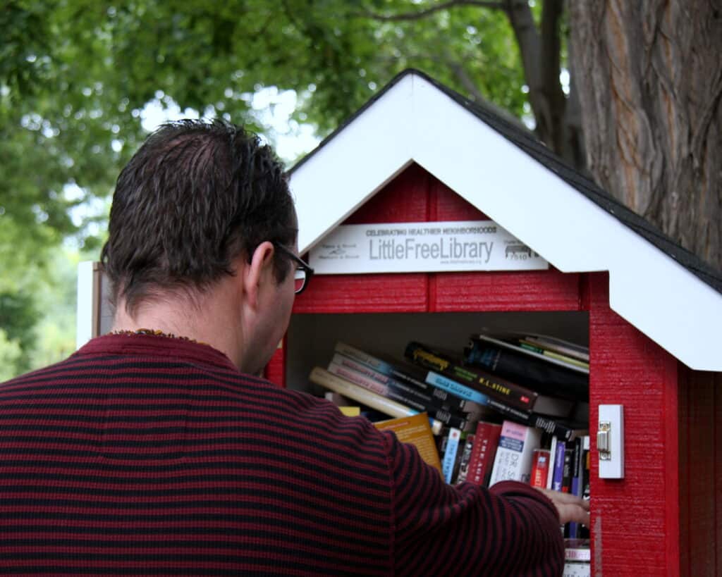 Books in public LittleFreeLibrary with a person browsing, outdoor setting with trees.