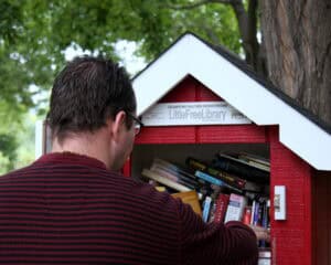 Books in public LittleFreeLibrary with a person browsing, outdoor setting with trees.