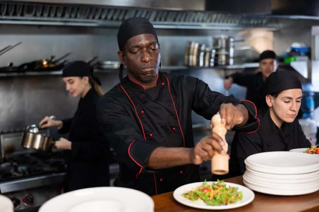 Confident african american chef in black uniform peppers salad in restaurant kitchen