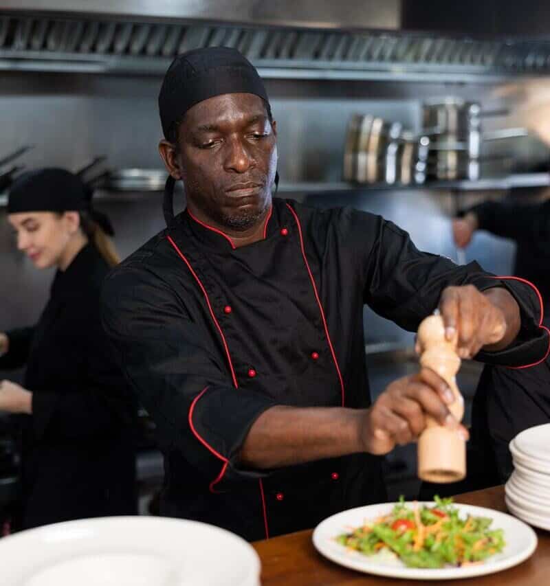 Confident african american chef in black uniform peppers salad in restaurant kitchen