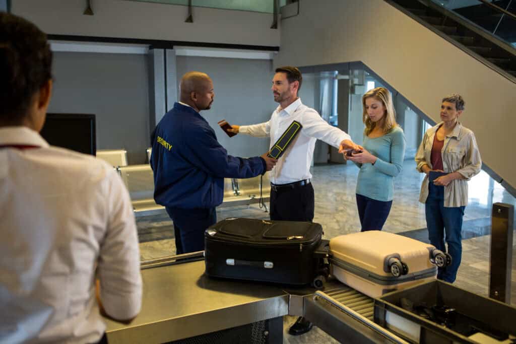 Airport security officers, assisted by a sniffer dog, inspect an open suitcase at a customs checkpoint while a passenger waits.