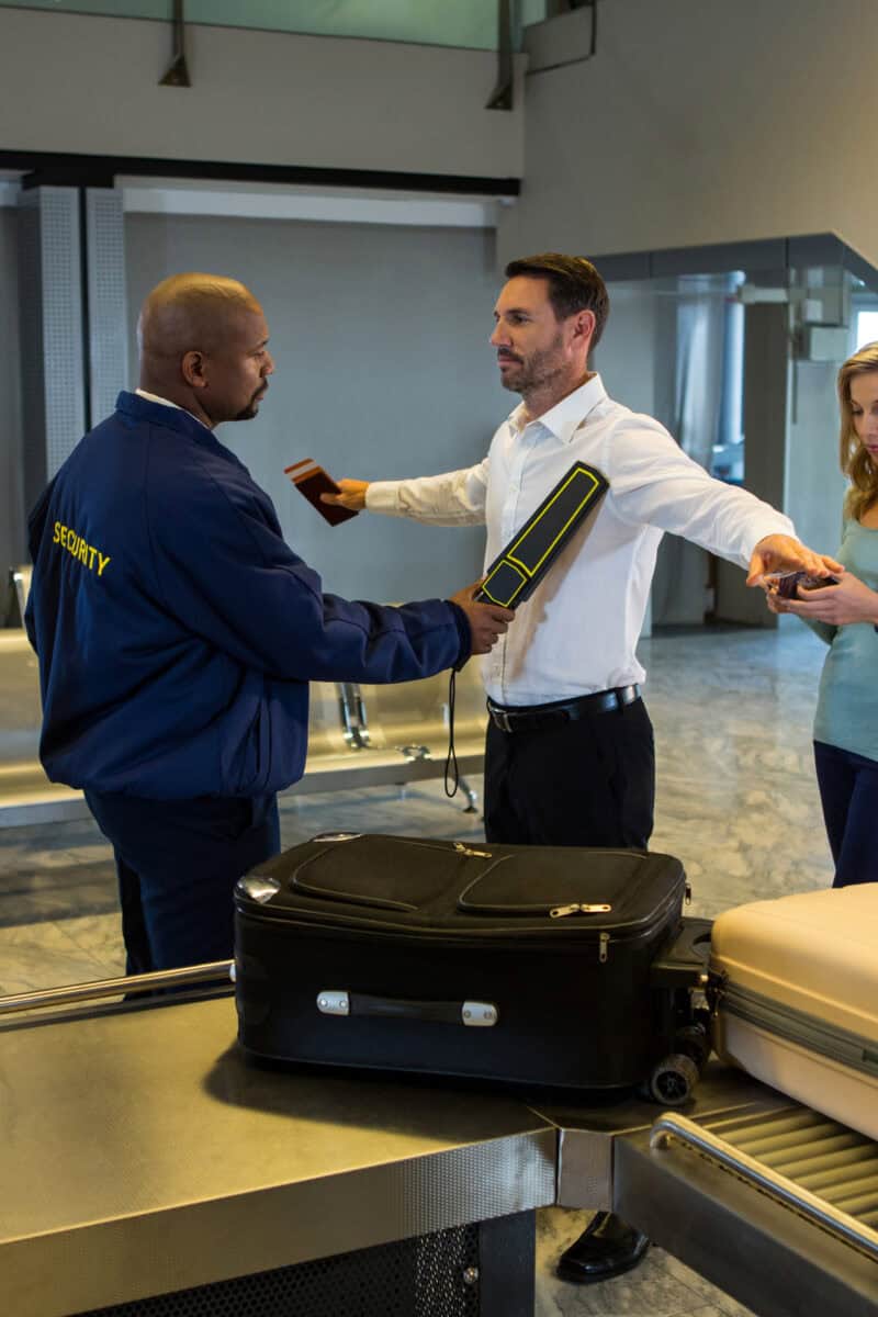 Airport security officers, assisted by a sniffer dog, inspect an open suitcase at a customs checkpoint while a passenger waits.