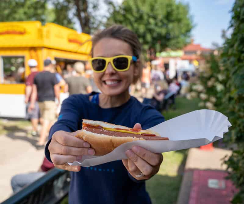 Smiling woman at an outdoor fair holds up a half-eaten footlong hot dog. Focus on the hot dog, girl intentionally blurred