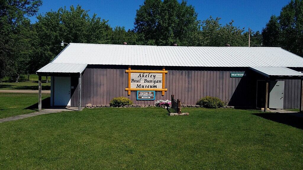 Akeley Paul Bunyan Museum building in a green park with clear blue sky, visitors information sign outside.