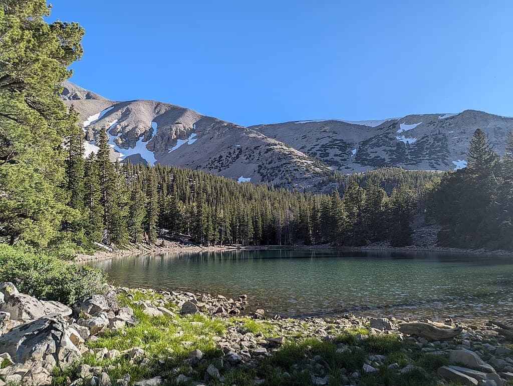 Serene mountain lake surrounded by pine trees and snow-capped peaks in clear blue sky.