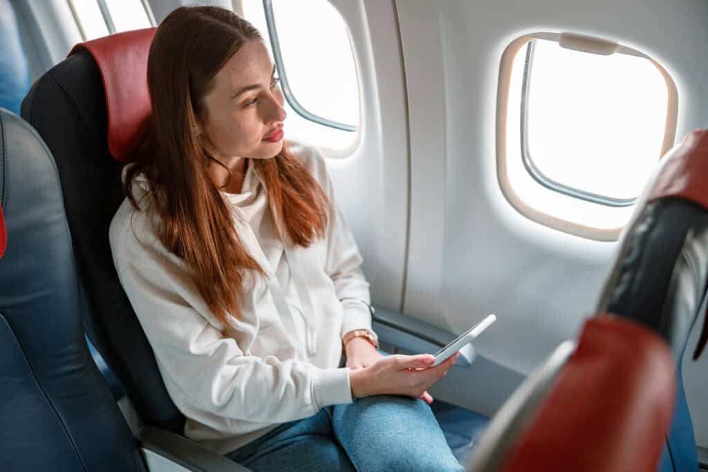 Female traveler with smartphone in her hand sitting in passenger chair and looking out the window in aircraft