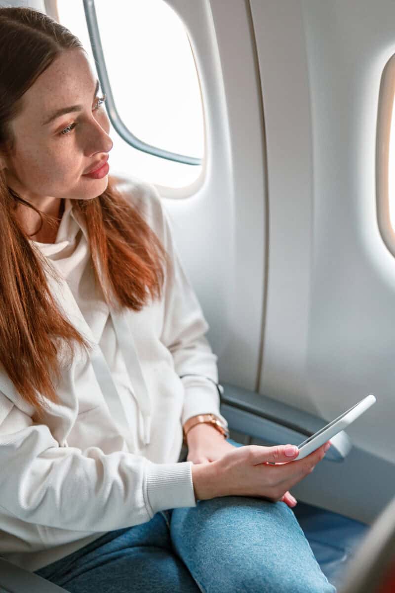 Female traveler with smartphone in her hand sitting in passenger chair and looking out the window in aircraft