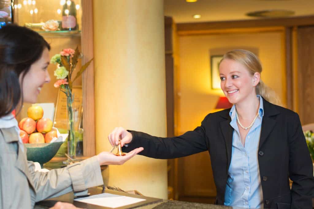 Smiling friendly hotel receptionist standing behind the service desk in a hotel lobby booking in a female client handing her the room keys for her stay during her vacation