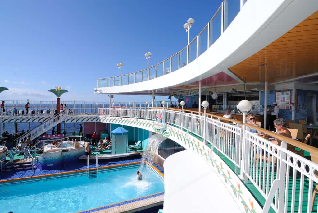 Seaside pool area on a cruise ship with outdoor bars and lounging spaces, sunny weather and blue sky.