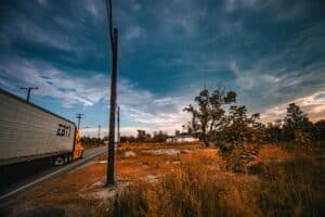 A scenic rural landscape featuring a trucking truck, wooden utility poles, and a vibrant sunset sky with scattered clouds.