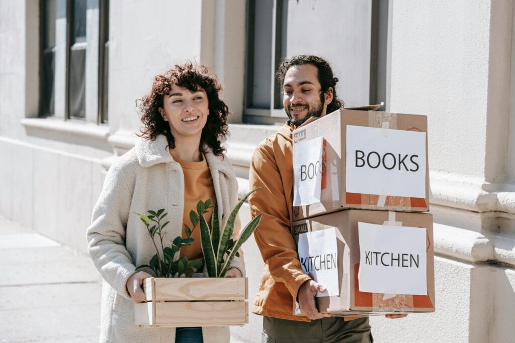 A Couple Moving Out Carrying Boxes And Plants