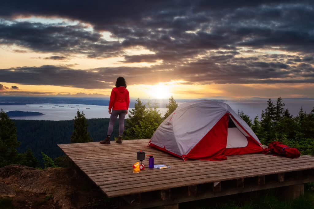 Adventure Girl and Camping Tent on top of a Mountain with Canadian Nature Landscape in the Background. Dramatic Morning Sunrise. Taken on Bowen Island, near Vancouver, British Columbia, Canada.