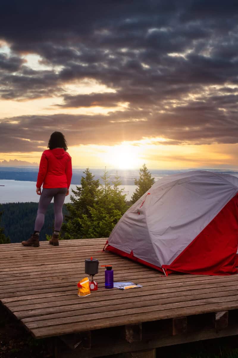 Adventure Girl and Camping Tent on top of a Mountain with Canadian Nature Landscape in the Background. Dramatic Morning Sunrise. Taken on Bowen Island, near Vancouver, British Columbia, Canada.