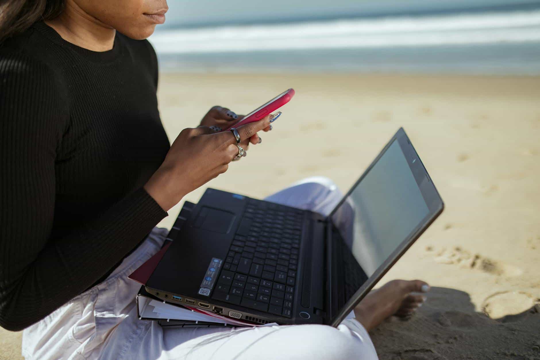 Person multitasking with a laptop and smartphone by the beach, embracing remote work lifestyle.