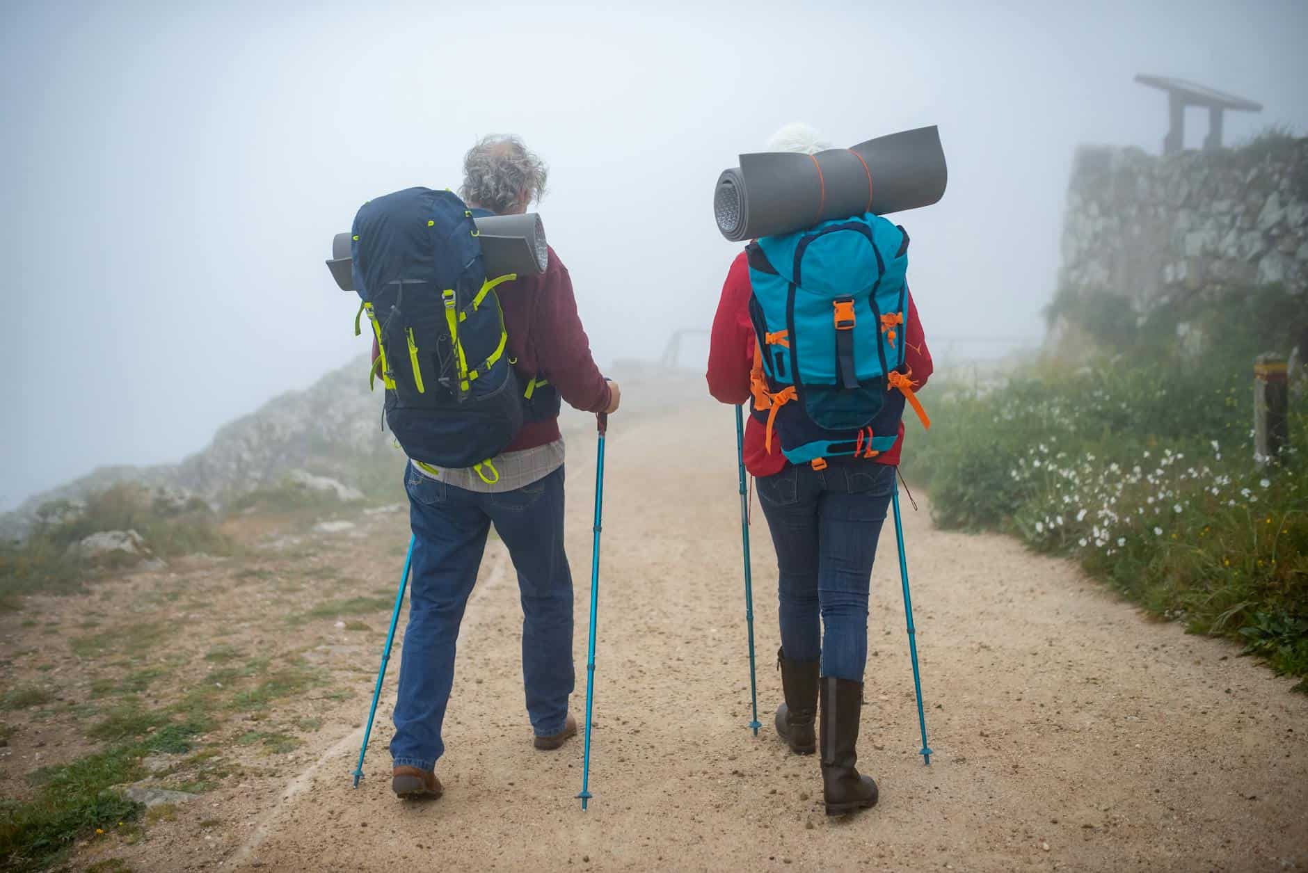 Elderly couple hiking on a foggy trail in Portugal, equipped with backpacks and walking poles.