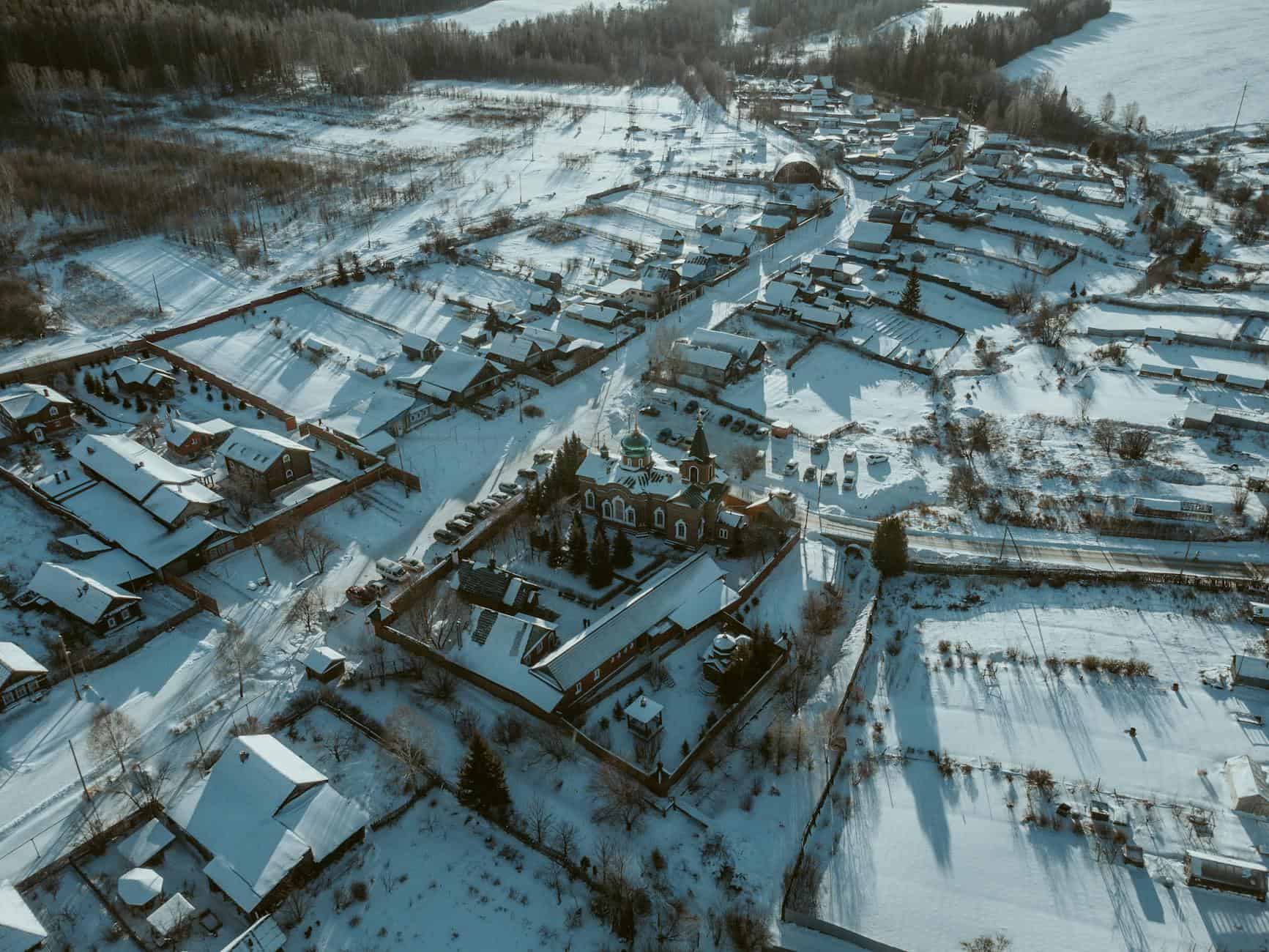 Aerial shot of a snow-covered rural village with scattered houses and a church surrounded by snowy fields.