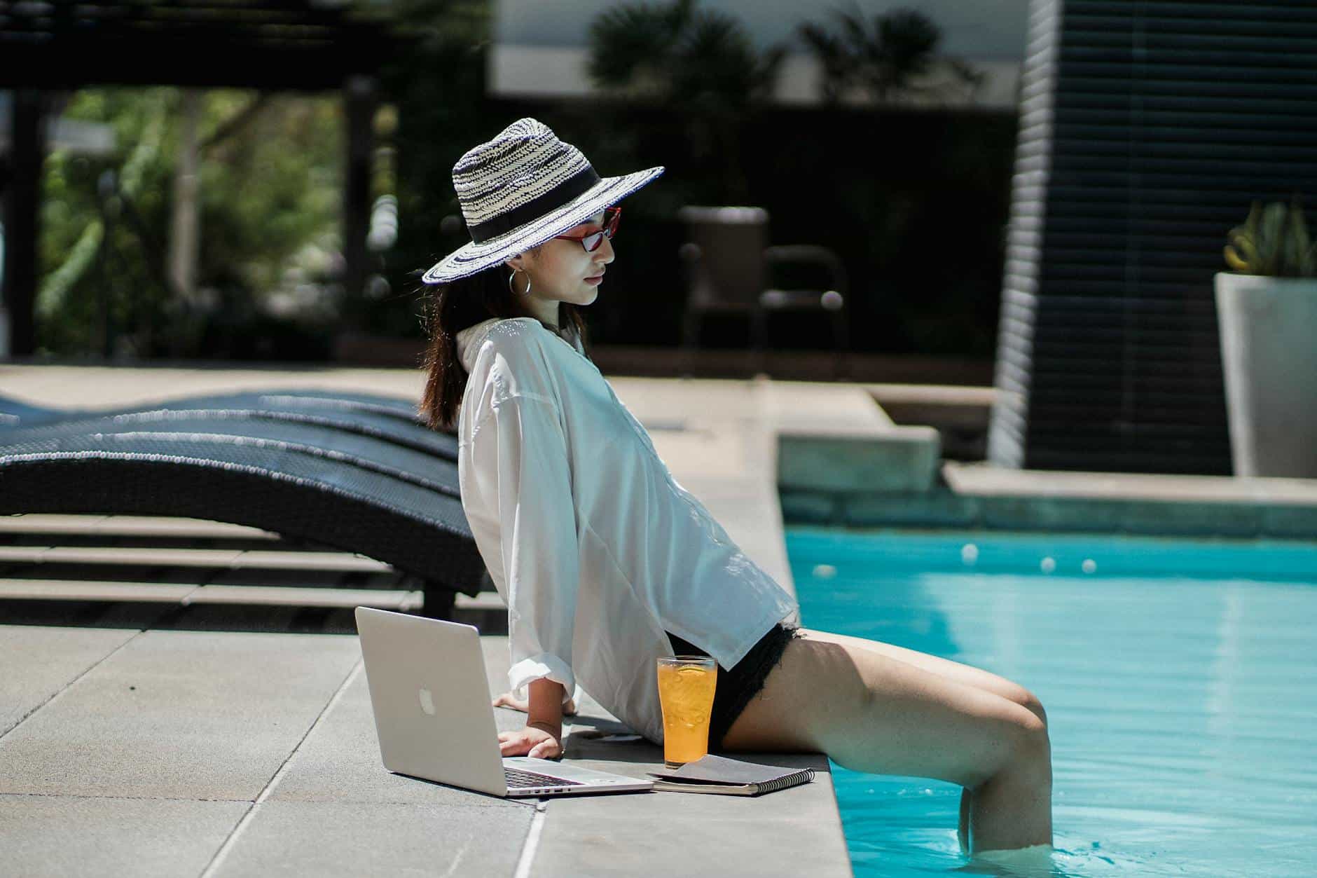 Side view of confident young Asian female freelancer in stylish clothes and hat sitting on outdoor swimming pool border with legs in water and resting after remote work on laptop