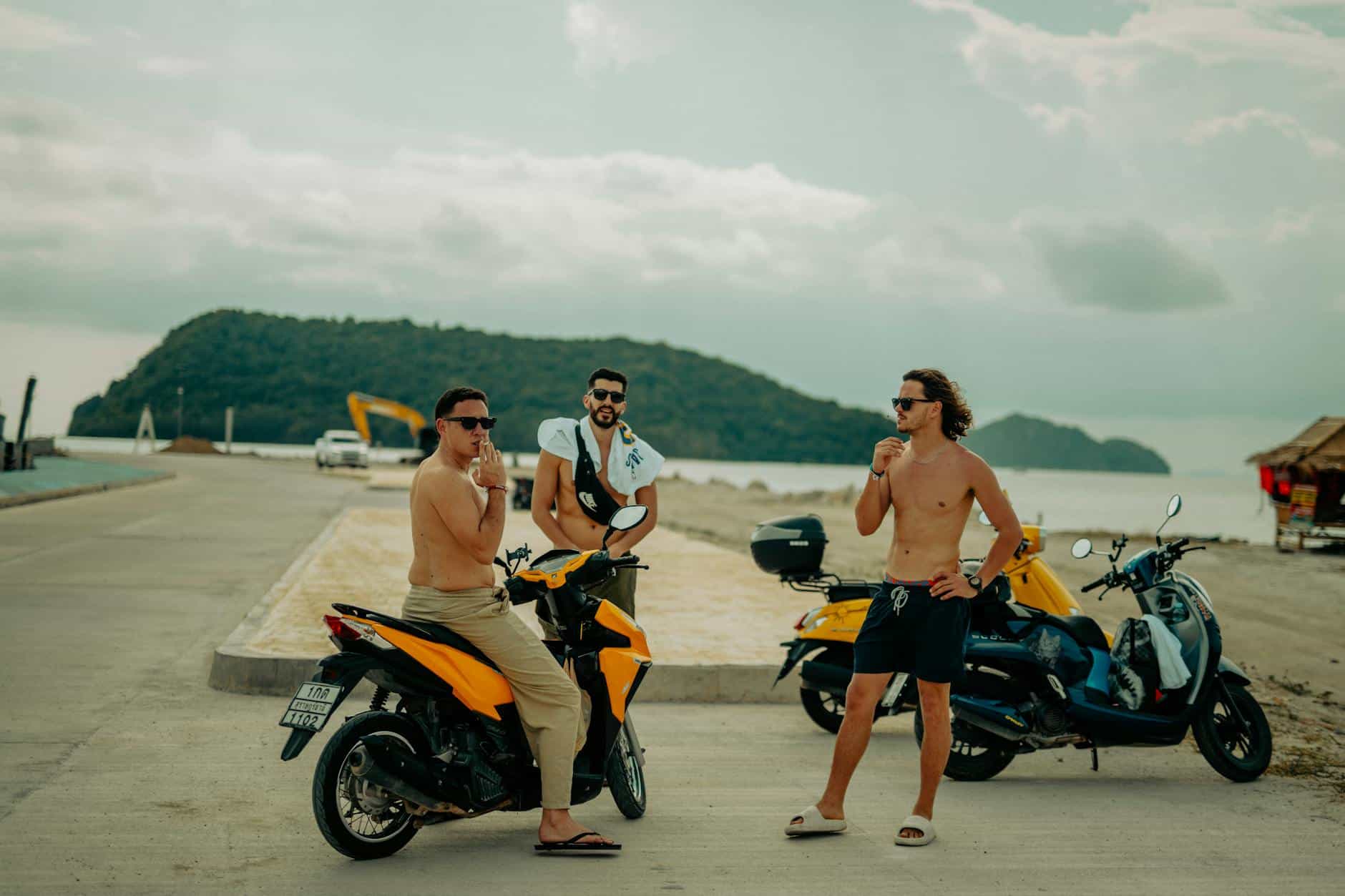 Group of friends enjoying a sunny day by the beach, leaning on scooters with a scenic island backdrop.