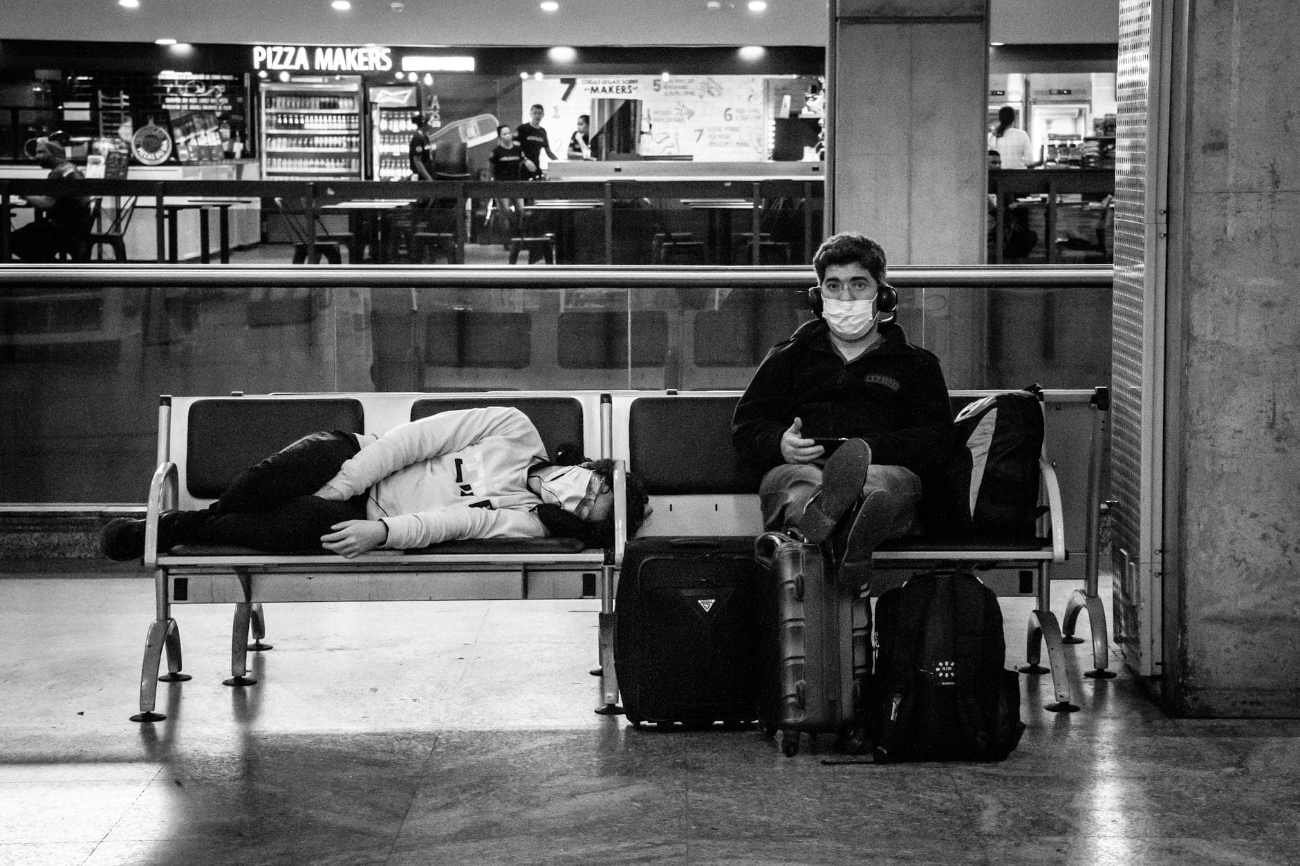 Black and white of unrecognizable woman sleeping on bench near sitting male traveler in sterile mask near suitcases in airport during quarantine period