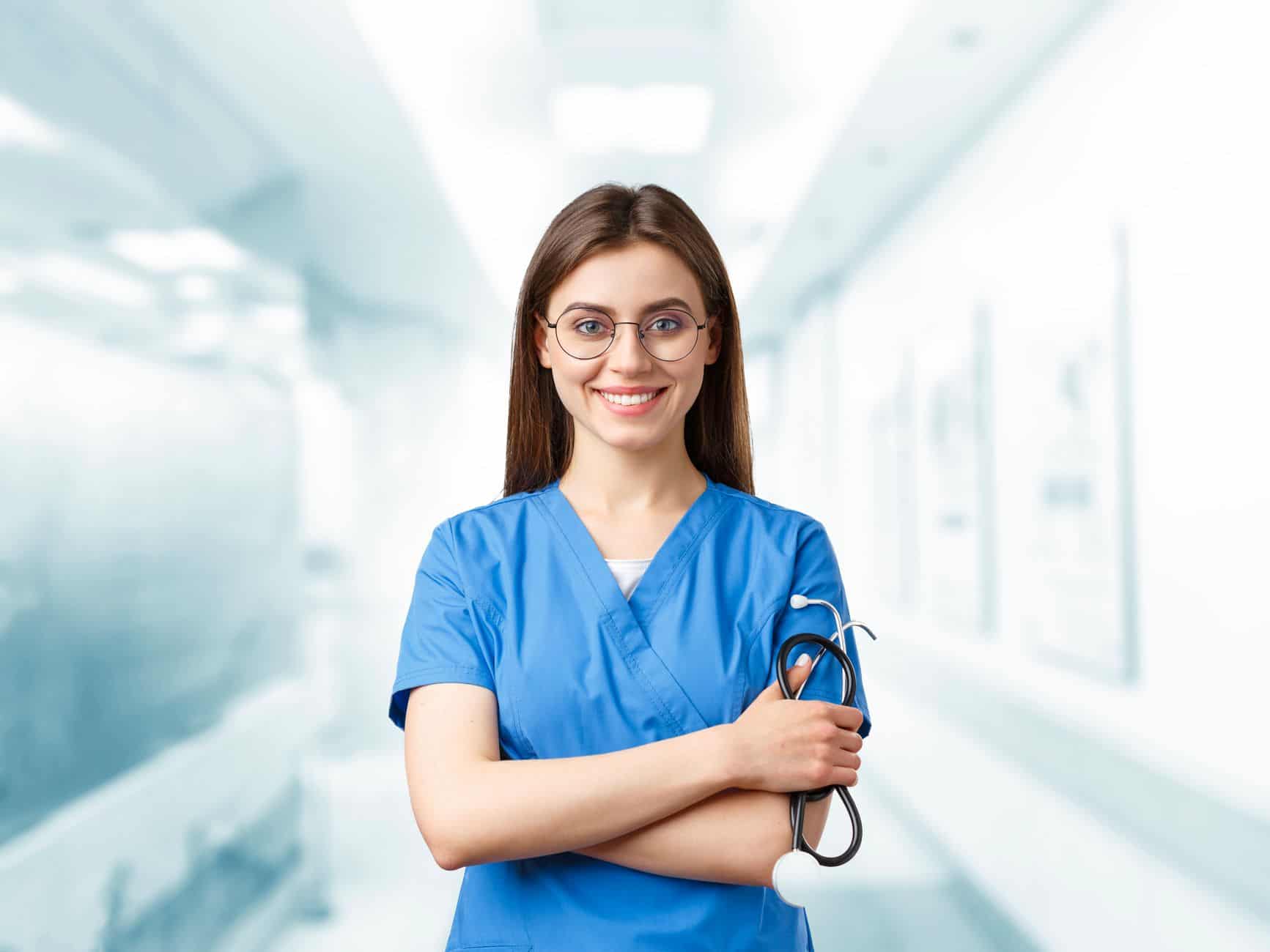 Young nurse in blue scrubs smiling and holding a stethoscope in a hospital corridor.