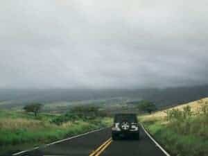 A jeep travels down a winding rural road surrounded by lush scenery under a cloudy sky.
