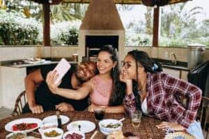 Three friends taking a selfie together at a table with food outdoors, capturing a joyful moment.