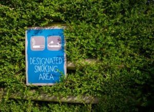Blue designated smoking area sign surrounded by lush greenery outdoors during the day.