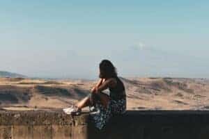 A woman sits on a wall enjoying the mountainous landscape and clear sky.