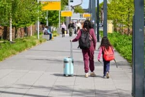 A mother and daughter walking with luggage outside Eindhoven Airport on a sunny day.