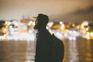 Man with backpack and hoodie standing by city lights at night. Perfect for travel and urban lifestyle themes.