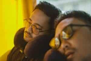 Two men wearing eyeglasses sleep on a bus, using neck pillows for comfort during the journey.
