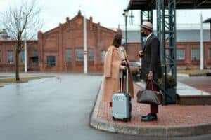 A fashionable couple in masks stands with luggage outside a historic building.