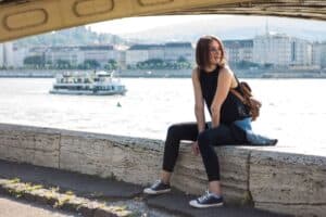 Casual young woman sitting by the riverside with cityscape and ferry in background.