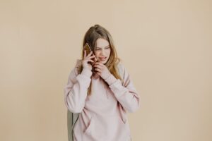 Casual young woman in pink hoodie talking on the phone against a beige background.