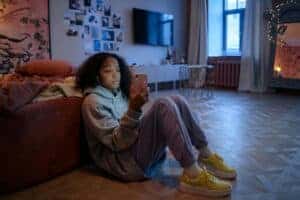 Teenage girl sitting on the floor using a smartphone, relaxed in a cozy room setting.