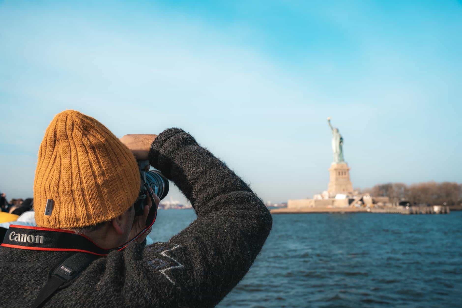 A person in a beanie capturing the Statue of Liberty across the water in New York City.