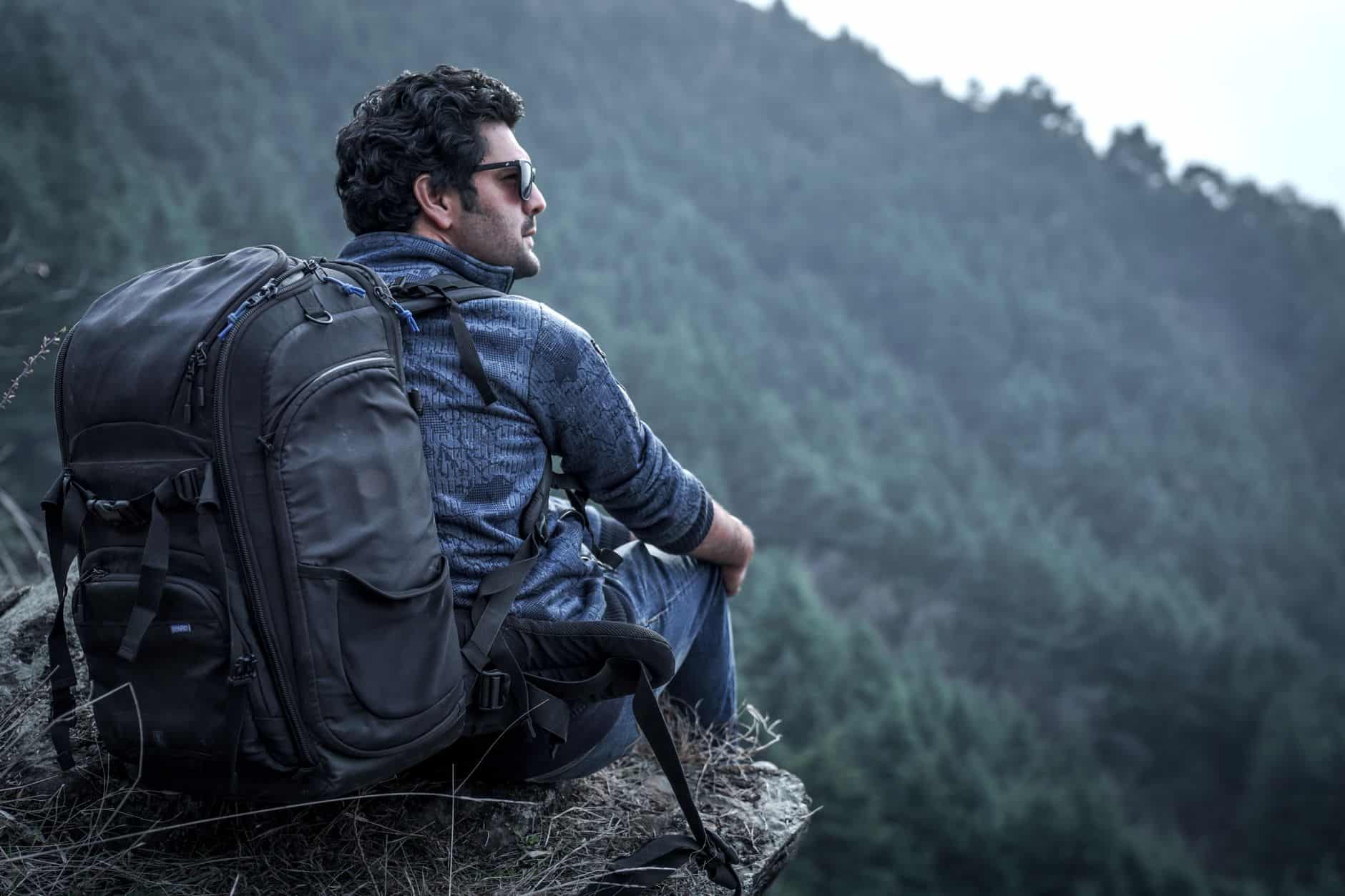 Backpacker sitting on cliff edge in forested mountains looking relaxed and adventurous.