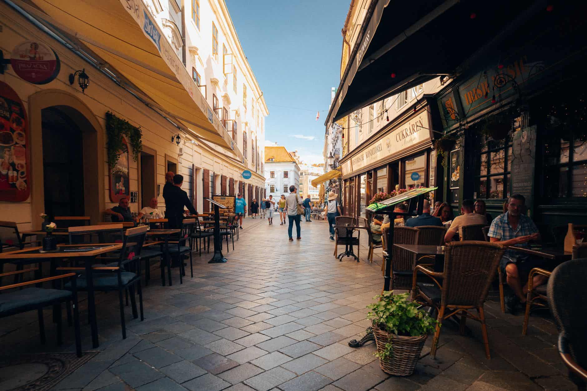 Lively café-lined street in Bratislava's historic city center under a clear sky.
