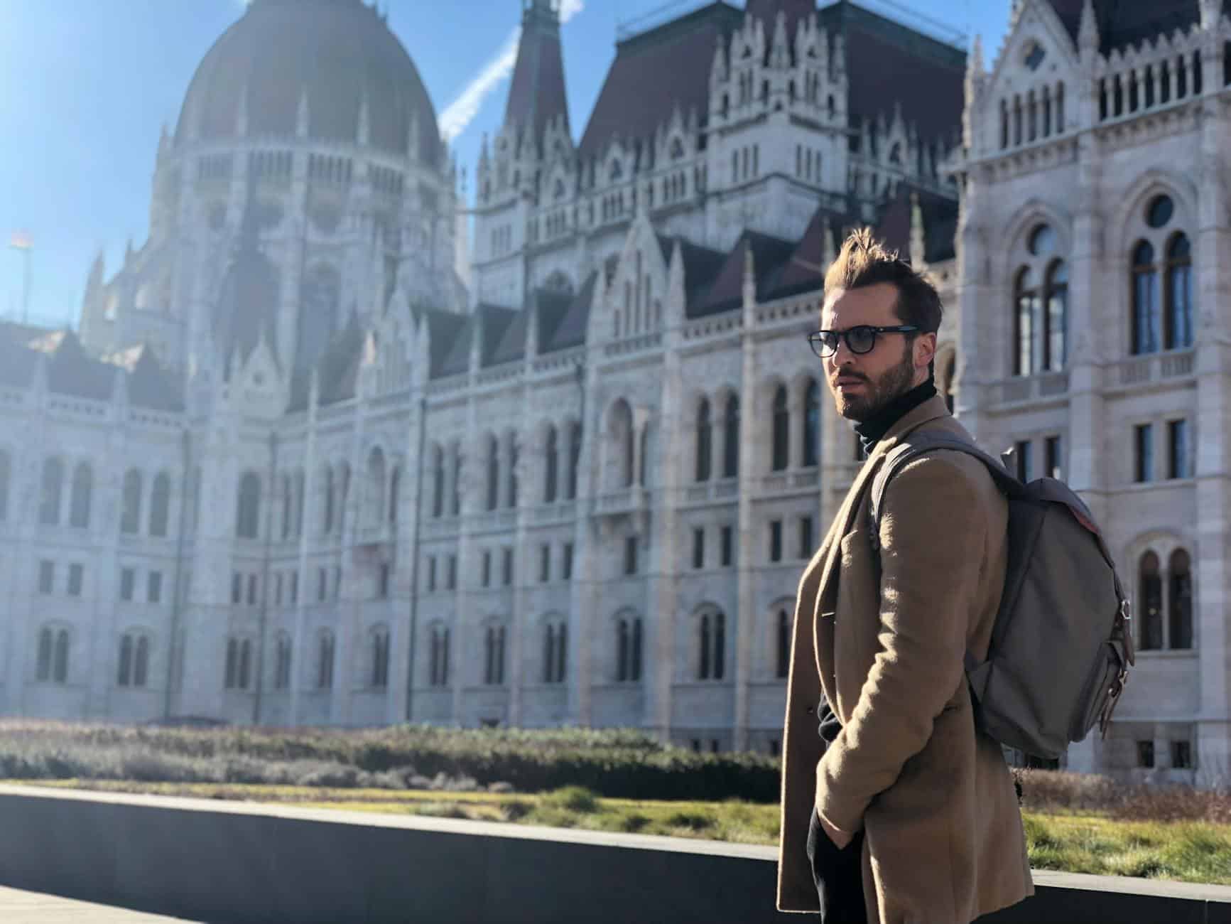 A man with a backpack stands in front of the historic Hungarian Parliament Building in Budapest.