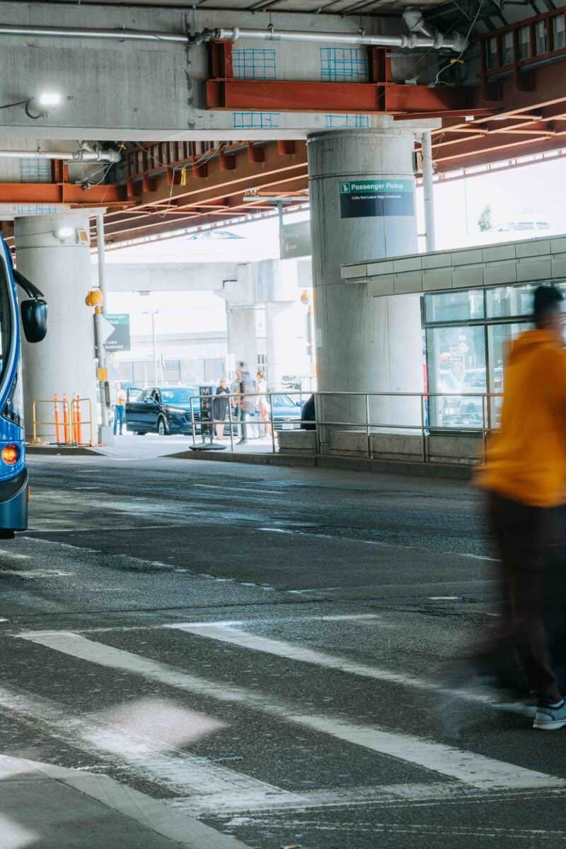 Busy airport scene showing travelers in motion and a rental car bus waiting under a covered terminal.