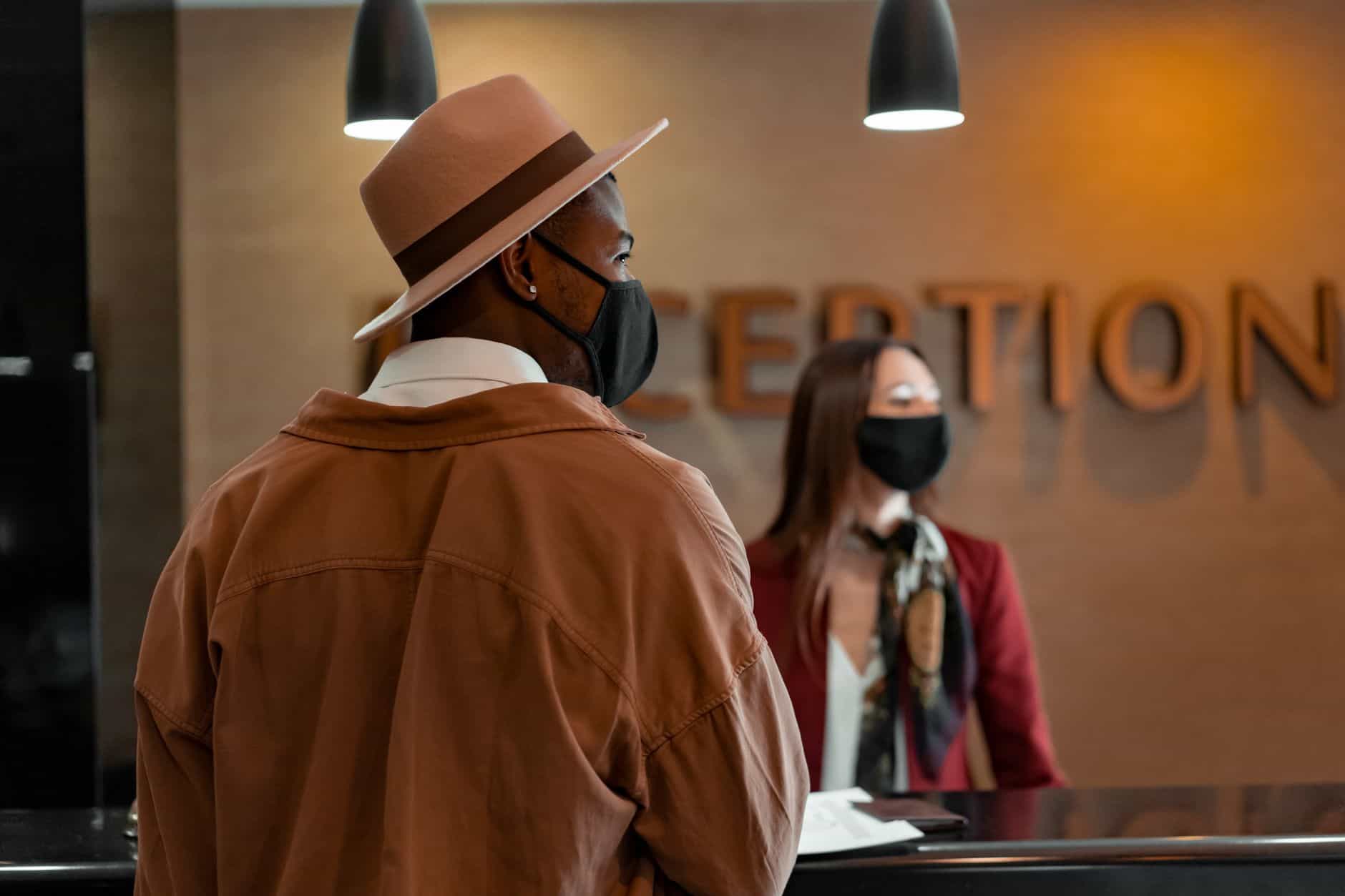 Man in mask at hotel reception during pandemic, interacting with masked receptionist.