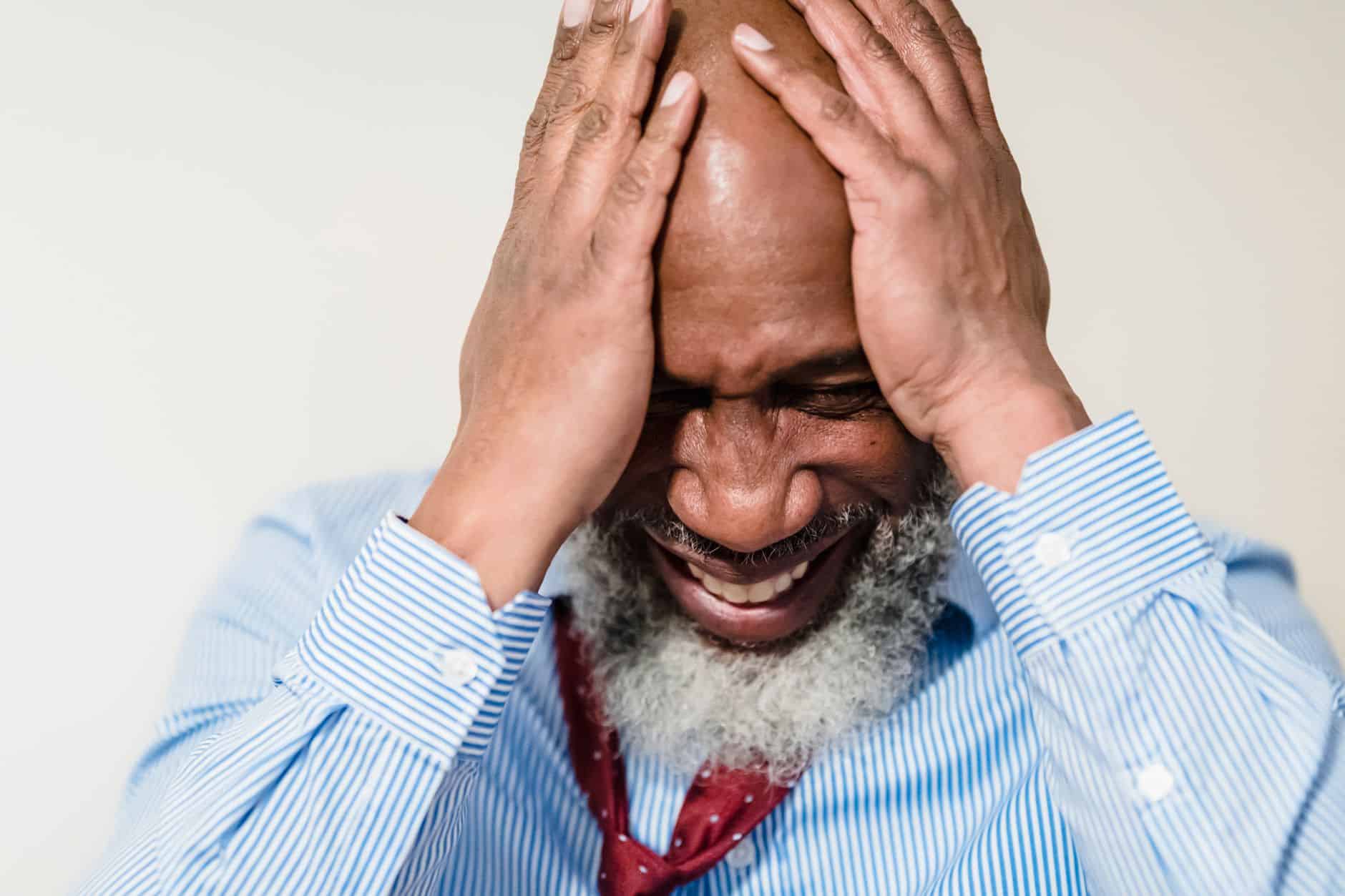 Elderly man in striped shirt showing stress and emotion with hands on head.