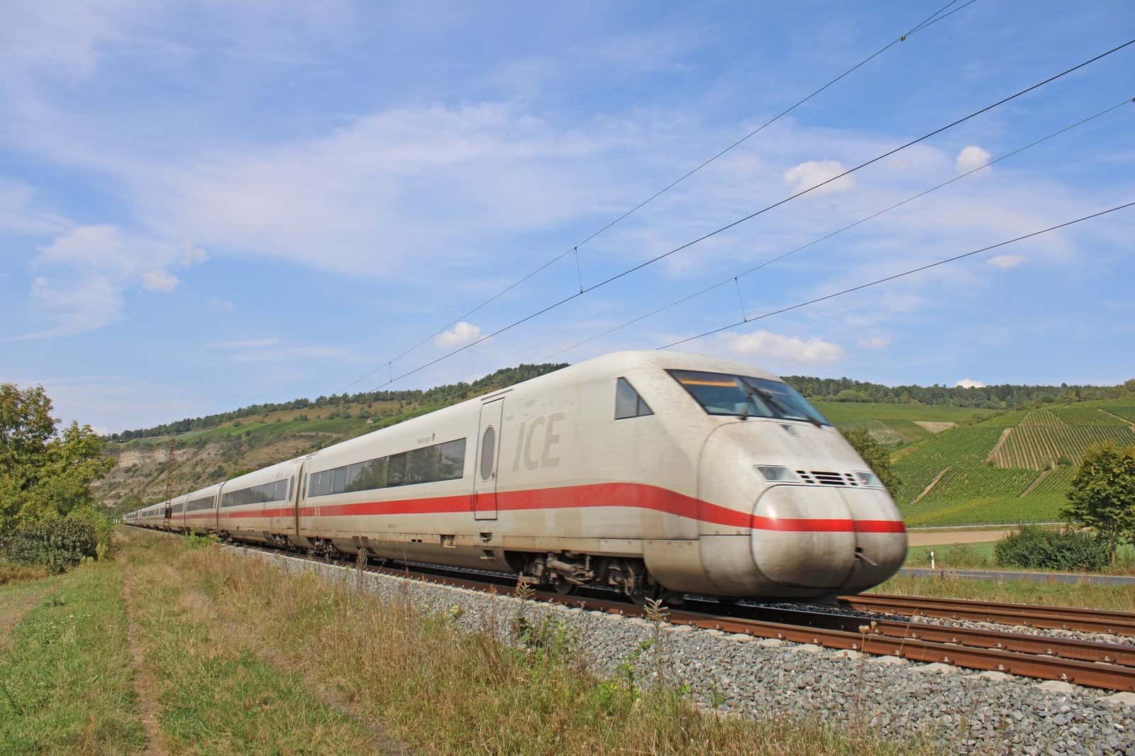 A high-speed train travels through a scenic rural countryside with green fields under blue skies.