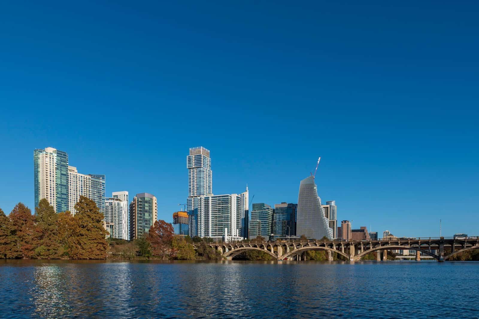 A vibrant city skyline with modern architecture reflecting in the river under clear blue skies.