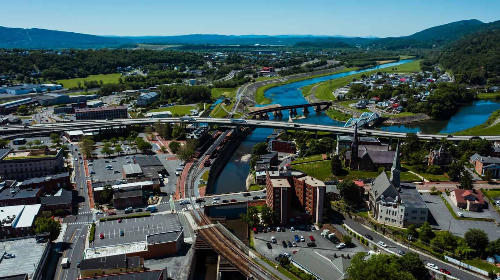 Aerial view of modern bridge with driving vehicles above canal and railroad behind mounts under blue sky in city